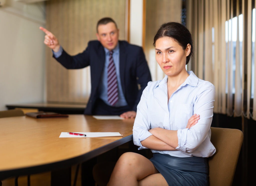 Frustrated woman entrepreneur sitting at office desk on background with angry partner expressing his dissatisfaction, pointing out mistakes in work